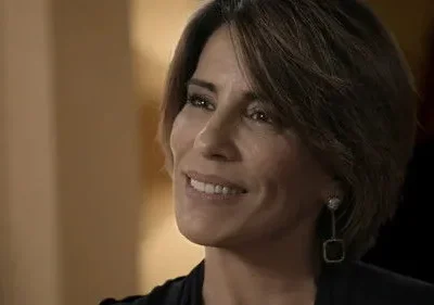 Mulher de cabelo curto sorri, olhando gentilmente para o lado. Fundo interno desfocado com tons quentes e suaves.