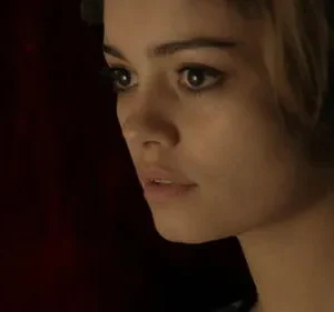 Close-up de mulher jovem, cabelo castanho e olhar intenso para o lado. Fundo escuro avermelhado, atmosfera sombria.