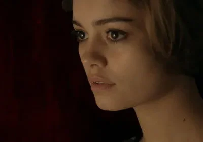 Close-up de mulher jovem, cabelo castanho e olhar intenso para o lado. Fundo escuro avermelhado, atmosfera sombria.