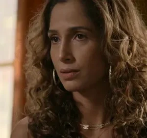 Close-up de mulher com cabelo cacheado e brincos de argola, com expressão pensativa. Fundo interno desfocado e iluminado.