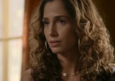 Close-up de mulher com cabelo cacheado e brincos de argola, com expressão pensativa. Fundo interno desfocado e iluminado.