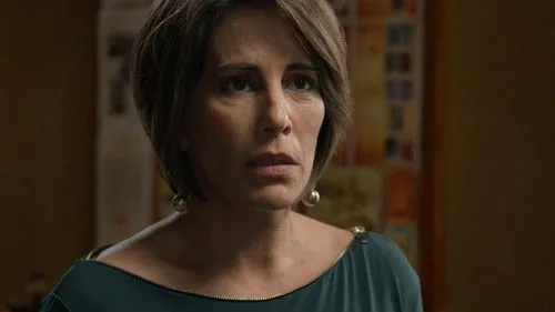 Babilônia – T01E60:Capítulo 60 1 Mulher de meia-idade com cabelo castanho curto,expressão preocupada,blusa verde escura e brincos dourados. Fundo embaçado.