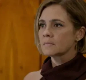 Mulher loira de cabelo curto, olhar sério e fixo, veste top vinho e brinco dourado. Fundo de madeira com veios.