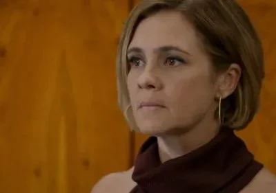 Mulher loira de cabelo curto, olhar sério e fixo, veste top vinho e brinco dourado. Fundo de madeira com veios.