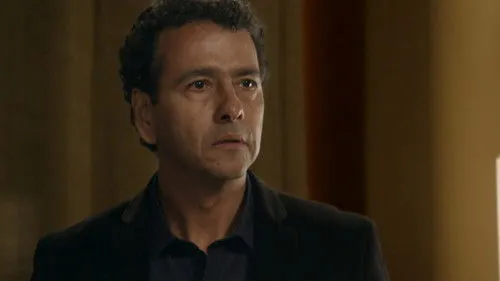 Babilônia – T01E62:Capítulo 62 1 Close-up de homem de meia-idade com cabelo escuro,olhando com expressão séria. Veste camisa e blazer escuros. Fundo em tons quentes e desfocado.