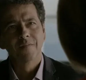 Homem de meia-idade, cabelo escuro e sorriso sutil, veste paletó e olha para o lado em ambiente com luz suave.