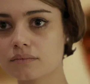 Mulher de cabelo bob castanho e olhar fixo. Uma figura borrada em segundo plano. Clima sério e enigmático.