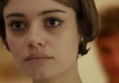 Mulher de cabelo bob castanho e olhar fixo. Uma figura borrada em segundo plano. Clima sério e enigmático.