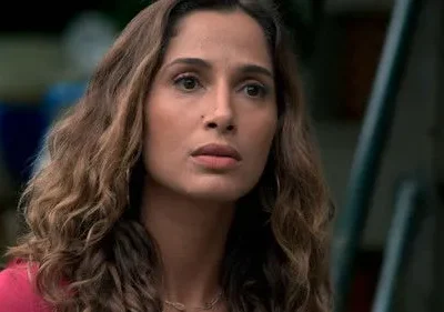 Mulher de cabelo castanho ondulado e top rosa, com expressão séria e pensativa, olhando para o lado. Fundo urbano desfocado.