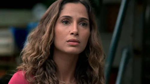 Babilônia – T01E70:Capítulo 70 1 Mulher de cabelo castanho ondulado e top rosa,com expressão séria e pensativa,olhando para o lado. Fundo urbano desfocado.