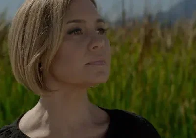 Mulher loira de cabelo curto olha pensativa para o alto em um campo de grama alta. Clima sereno e natural.