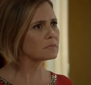 Mulher de cabelos curtos loiros, expressão séria, vestindo blusa vermelha com detalhes prateados, em ambiente interno. Clima pensativo.