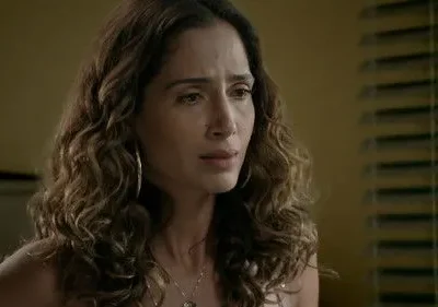 Mulher de cabelo encaracolado e brincos, com expressão melancólica. Fundo com parede amarela e persianas.