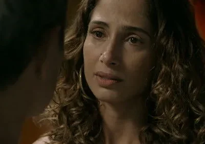 Mulher de cabelos cacheados com expressão séria olha para uma pessoa com o ombro visível. Fundo escuro avermelhado.