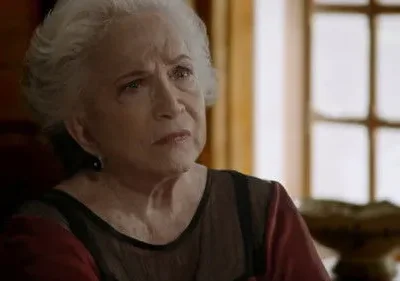 Mulher idosa de cabelo branco e expressão séria. Em um ambiente interno, com janela e detalhes de madeira no fundo desfocado.