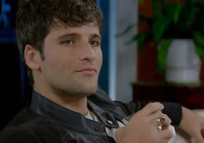 Jovem de cabelo escuro e olhos azuis, casaco preto. Ele sorri levemente e segura um objeto com anel vistoso. Fundo interno com planta.