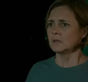 Mulher de cabelo curto e rosto tenso, com olhar preocupado num ambiente escuro, banhado por uma luz verde misteriosa. Clima de suspense.