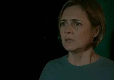 Mulher de cabelo curto e rosto tenso, com olhar preocupado num ambiente escuro, banhado por uma luz verde misteriosa. Clima de suspense.