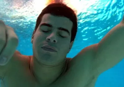 Homem de olhos fechados submerso em água azul clara. Um braço estendido, outro com punho cerrado. Luz do sol na superfície.