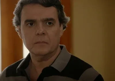 Homem de meia-idade com cabelo grisalho e olhar arregalado, preocupado. Veste camisa polo listrada, fundo desfocado.