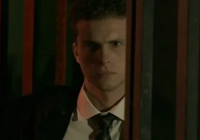 Homem sério de terno atrás de barras escuras, iluminado dramaticamente. O clima é tenso e misterioso.