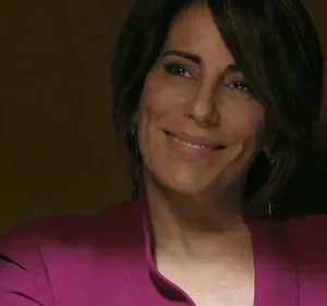 Mulher de cabelo castanho e blazer magenta sorri gentilmente. Fundo escuro e discreto, com um toque de elegância no ambiente.