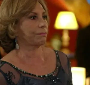 Mulher de cabelo loiro em vestido de festa azul escuro, com expressão séria, em um ambiente formal com iluminação quente e figuras desfocadas.