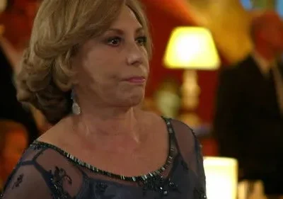 Mulher de cabelo loiro em vestido de festa azul escuro, com expressão séria, em um ambiente formal com iluminação quente e figuras desfocadas.