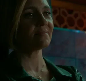 Mulher loira em jaqueta escura, olhar pensativo em ambiente escuro. Fundo com padrões e luzes em tons quentes e frios.