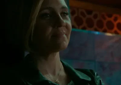 Mulher loira em jaqueta escura, olhar pensativo em ambiente escuro. Fundo com padrões e luzes em tons quentes e frios.