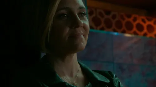 Babilônia – T01E96:Capítulo 96 1 Mulher loira em jaqueta escura,olhar pensativo em ambiente escuro. Fundo com padrões e luzes em tons quentes e frios.