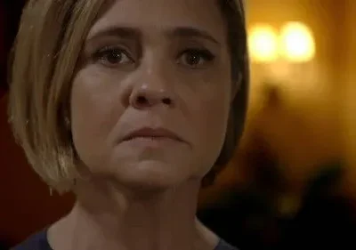 Close-up de mulher com cabelo curto e expressão sombria, olhando fixamente. Fundo escuro com luz ambiente quente e borrada.