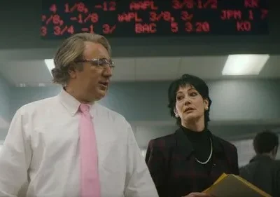 Um homem de gravata rosa fala com uma mulher atenta em um escritório. Painel de cotações de ações com números vermelhos ao fundo.