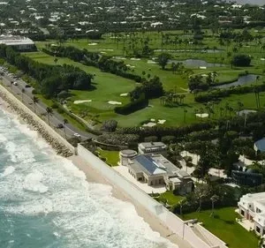 Vista aérea de costa luxuosa: oceano, praia, mansões, e um vasto campo de golfe com palmeiras e lagos.