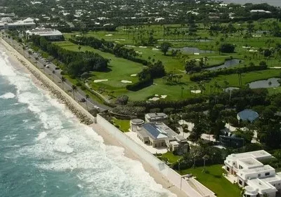 Vista aérea de costa luxuosa: oceano, praia, mansões, e um vasto campo de golfe com palmeiras e lagos.