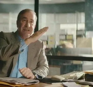 Um homem de terno, com expressão animada, gesticula com a mão aberta em uma mesa de escritório. Janelas ao fundo revelam um cenário urbano.