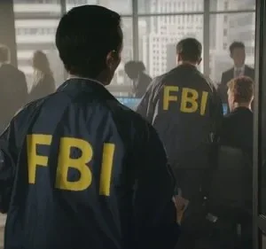 Agentes do FBI, de costas, usam jaquetas azuis com FBI amarelo. Estão em um escritório com vista para a cidade. Ambiente sério.