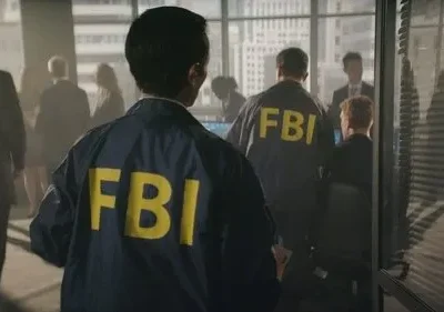 Agentes do FBI, de costas, usam jaquetas azuis com FBI amarelo. Estão em um escritório com vista para a cidade. Ambiente sério.