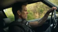 Homem de perfil, com colete tático e marcas no pescoço, dirige um carro. Olha sério para a frente, com árvores borradas ao fundo.