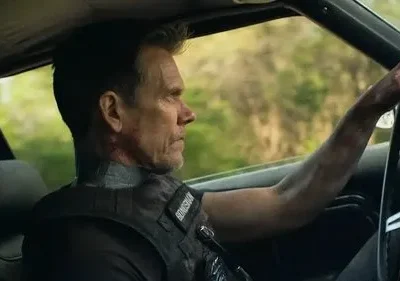 Homem de perfil, com colete tático e marcas no pescoço, dirige um carro. Olha sério para a frente, com árvores borradas ao fundo.