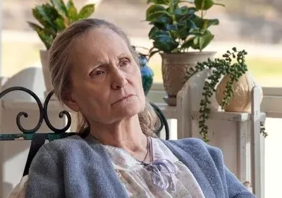 Mulher idosa séria de cabelos claros e cardigã azul, sentada em varanda. Plantas em vasos ao fundo criam um clima tranquilo.