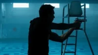 Homem em silhueta mira arma em piscina escura e azulada com cadeira de salva-vidas. Ambiente misterioso e tenso.