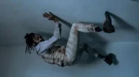 Homem de dreadlocks e terno claro sujo está suspenso, braços erguidos, em um cômodo simples e escuro. Imagem surreal.