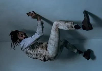 Homem de dreadlocks e terno claro sujo está suspenso, braços erguidos, em um cômodo simples e escuro. Imagem surreal.
