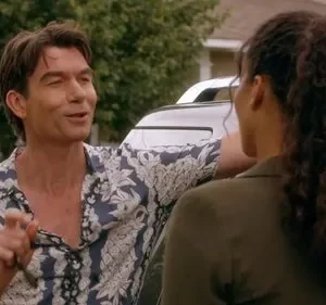 Homem de camisa floral azul sorri e gesticula para mulher de cabelo cacheado, vista de costas. Ambos encostados em carro, rua residencial.