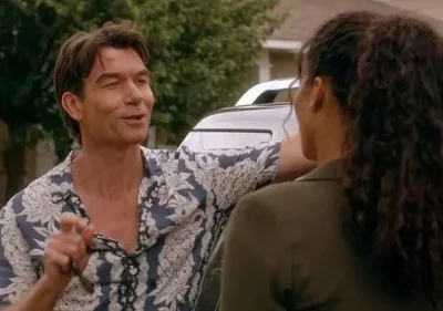 Homem de camisa floral azul sorri e gesticula para mulher de cabelo cacheado, vista de costas. Ambos encostados em carro, rua residencial.