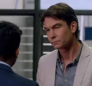 Um homem de blazer claro olha sério para as costas de outro homem de terno, em um ambiente interno bem iluminado.