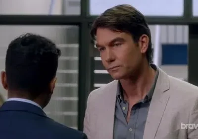 Um homem de blazer claro olha sério para as costas de outro homem de terno, em um ambiente interno bem iluminado.