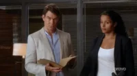 Homem de casaco claro e mulher de blazer em escritório. Ele olha sério, segurando documentos; ela observa atentamente. Clima profissional.