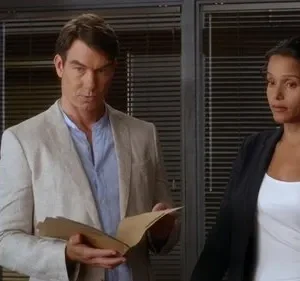 Homem de casaco claro e mulher de blazer em escritório. Ele olha sério, segurando documentos; ela observa atentamente. Clima profissional.